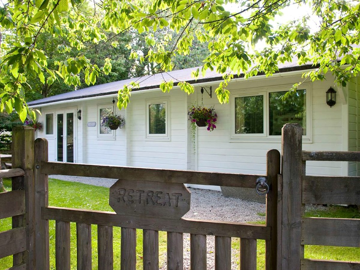 A long white single-storey lodge with a slate roof, hanging baskets and French doors, behind a wooden gate carved with the word Retreat, set on a lawn among trees.