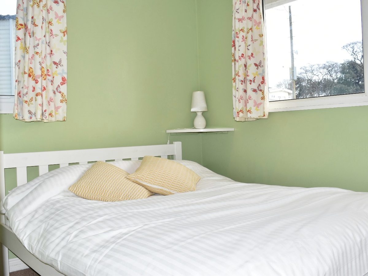 Double bedroom with a white wooden bed frame, white bedding and yellow cushions against a green wall, with floral curtains at the window.