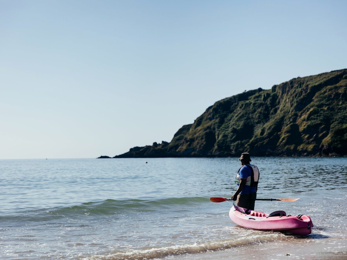 A person standing in the shallows beside a pink sit-on kayak pulled up at the water's edge, with a green headland behind.