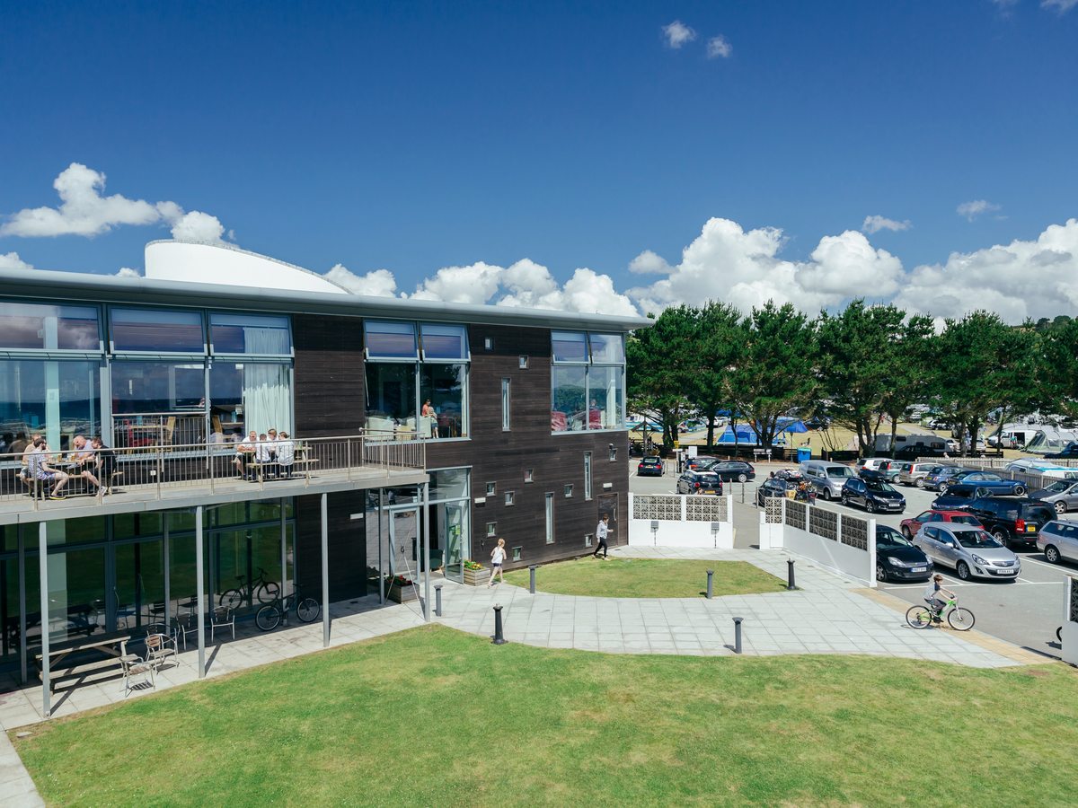 A two-storey timber-clad building with a glass frontage and a first-floor balcony where people sit at tables, a lawn and paved paths in front and a car park to one side.