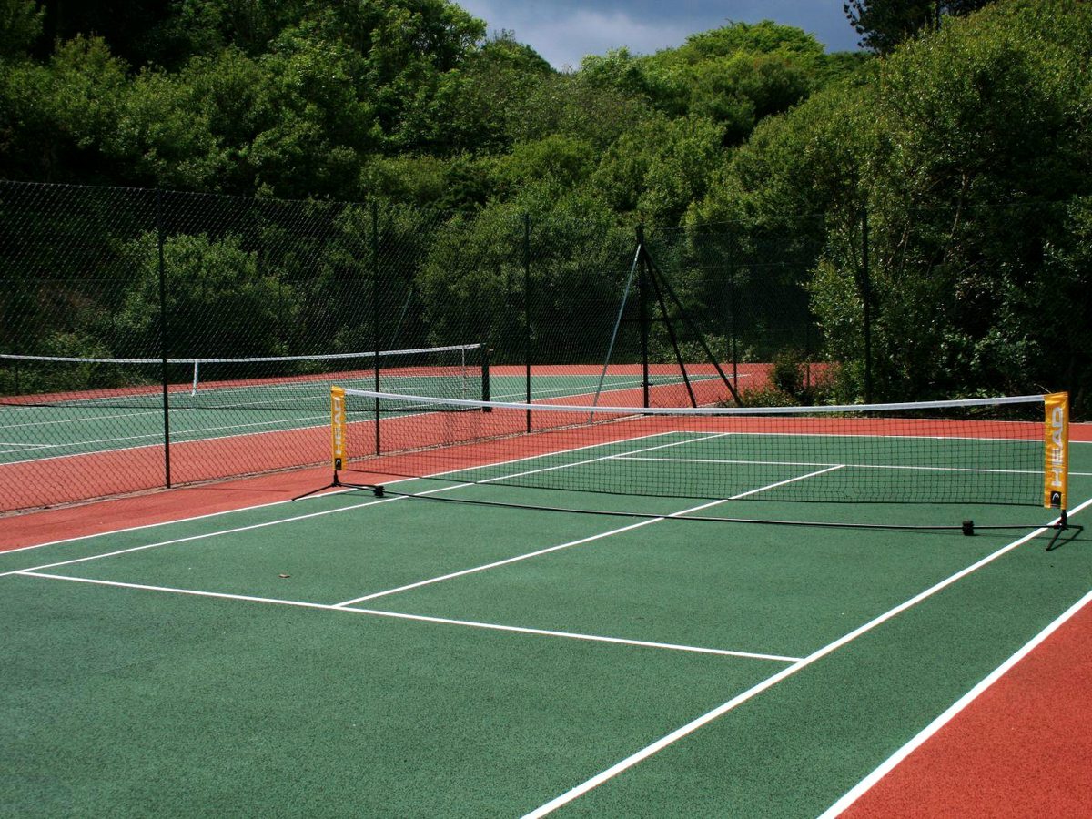 A fenced outdoor tennis court with a green playing surface and red surround, the net up, backed by trees.