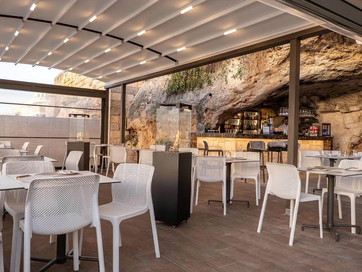 Terrace of white tables and chairs under a slatted canopy, with the bar counter set back beneath a natural rock overhang and a member of staff behind it.