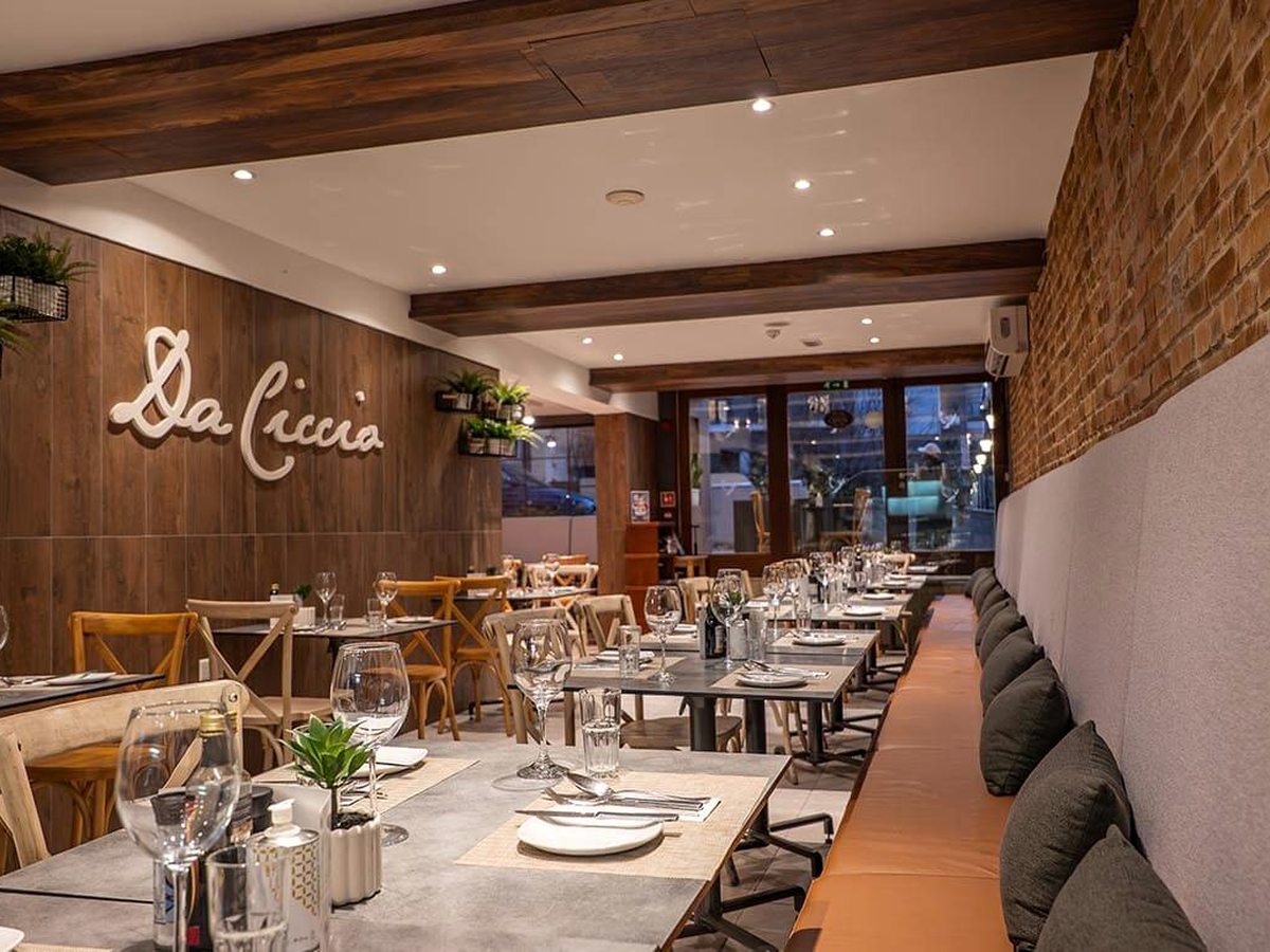 Restaurant interior with the Da Ciccio name in script on a timber-panelled wall, laid tables and wine glasses along a cushioned banquette, exposed brick to one side and dark ceiling beams.