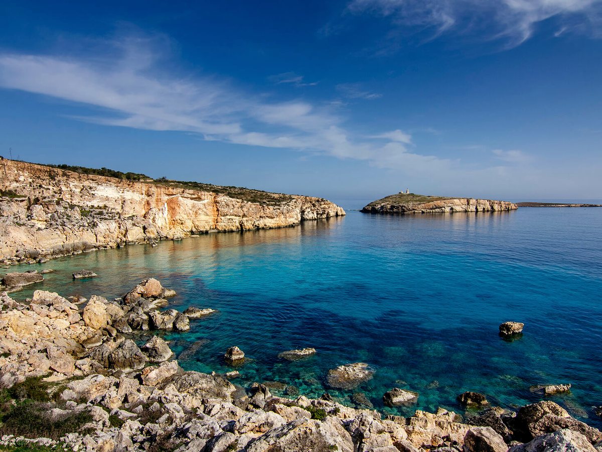 Clear turquoise water in a bay edged by pale limestone rocks, with a low headland and a small tower on the point under a bright blue sky.