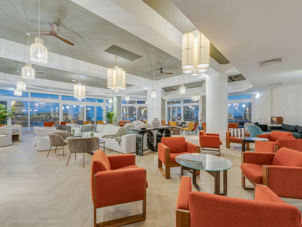 A wide open-plan hotel lobby lounge with burnt-orange armchairs, pale sofas, glass coffee tables and lantern pendant lights, with dusk showing through a wall of windows.