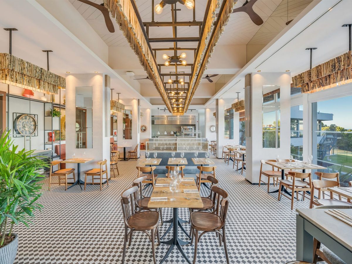 A restaurant with a chequerboard tiled floor, wooden bistro chairs, an open kitchen counter at the far end and rope-fringed light fittings overhead.