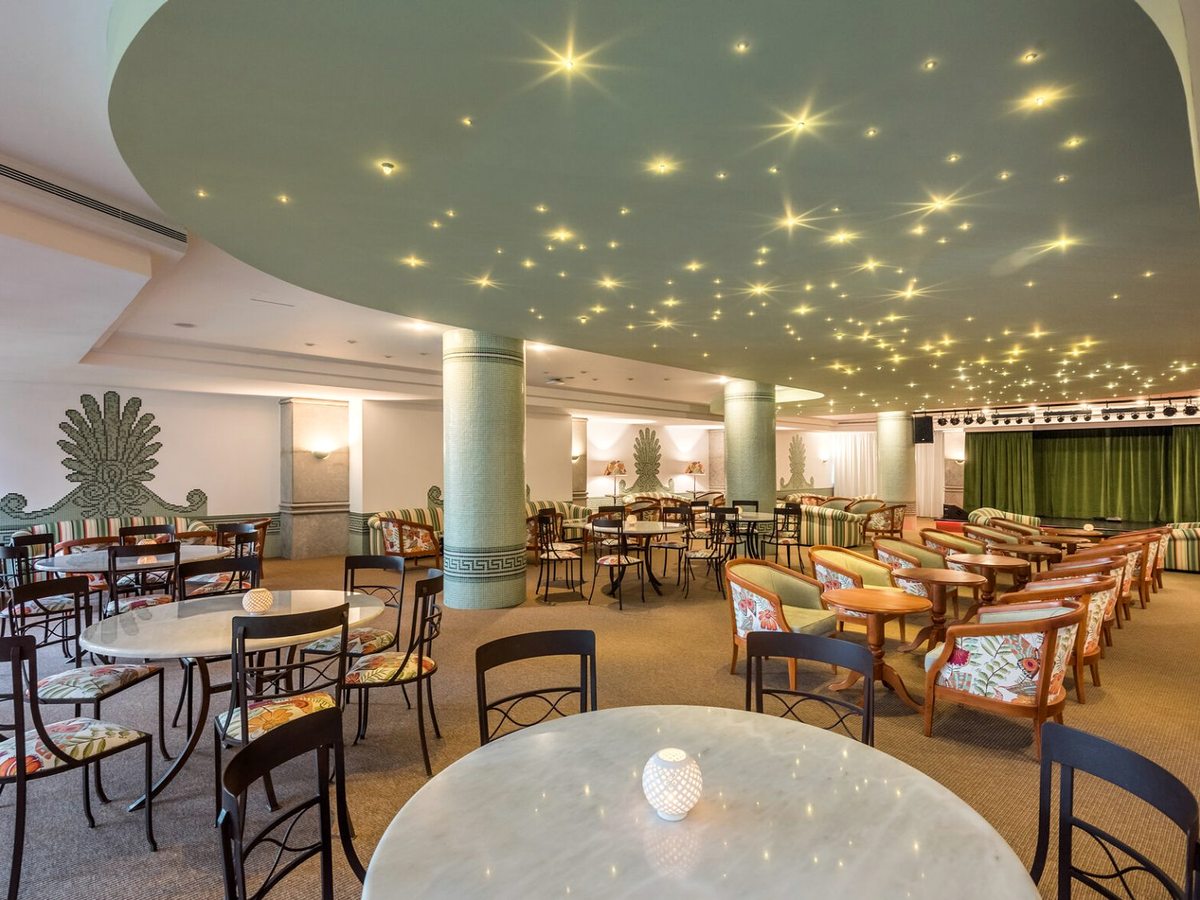 A large hotel lounge with marble-topped tables, floral armchairs, green mosaic columns, a domed ceiling of small star lights and a green-curtained stage at the far end.