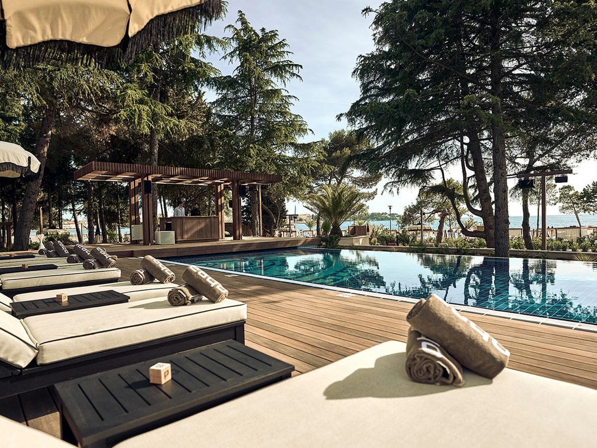 Rectangular pool edged with a timber deck and cushioned daybeds under tall pines, a covered bar pavilion beside it and the sea behind.