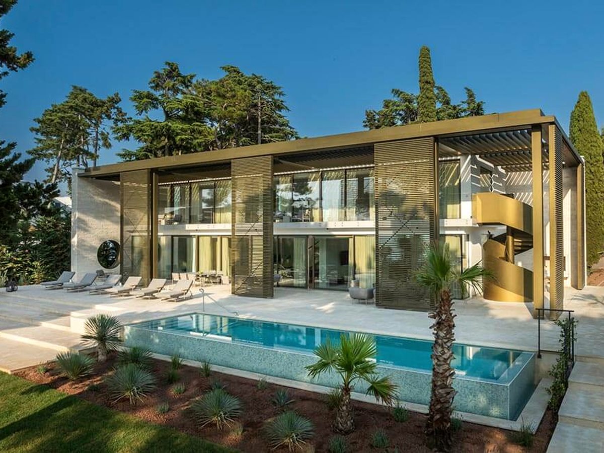 Two-storey glass and timber-louvred villa with a rectangular pool, sun loungers on a paved terrace and pines behind.