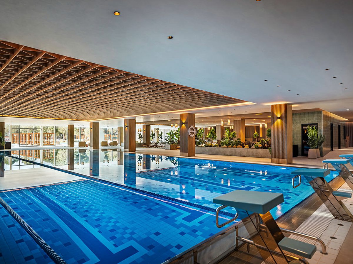 Indoor pool with lane markings and starting blocks along one edge, a timber-grid ceiling, timber-clad columns and loungers along the far side.