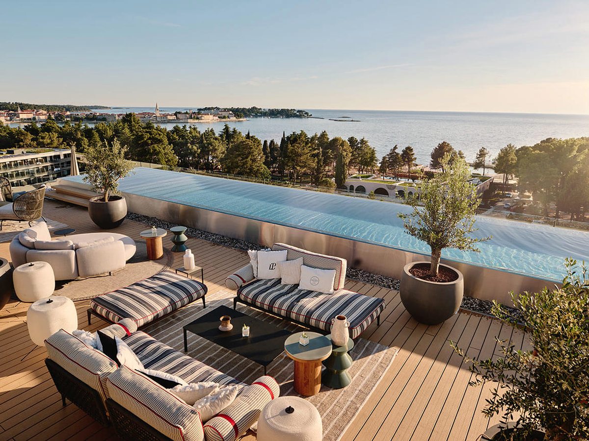 Rooftop terrace with striped daybeds, cream sofas and potted olive trees beside a long infinity pool, with the wooded coast and Poreč old town beyond.