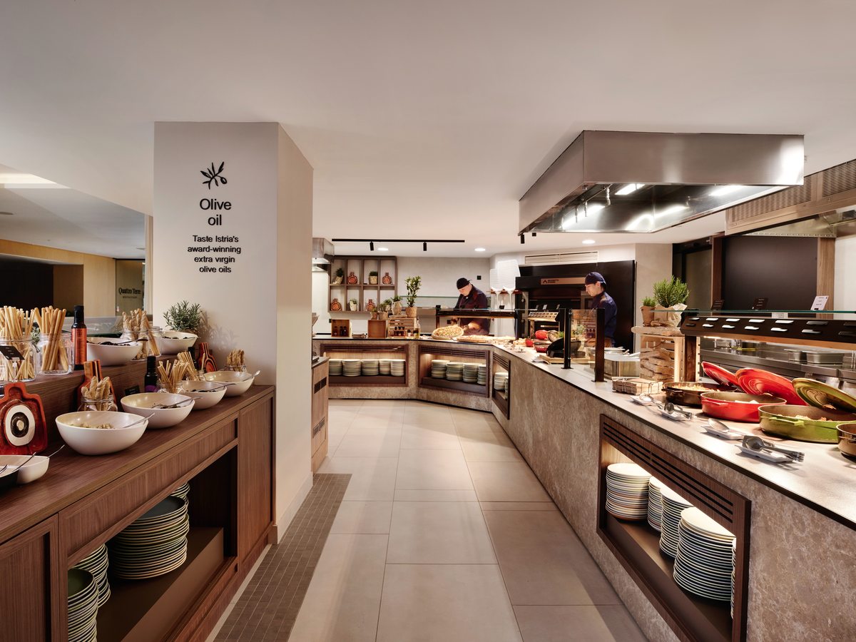 Buffet counters set with bowls, breadsticks and stacked plates, a hot service section opposite and chefs working at an open kitchen behind, with a pillar signed Olive oil.