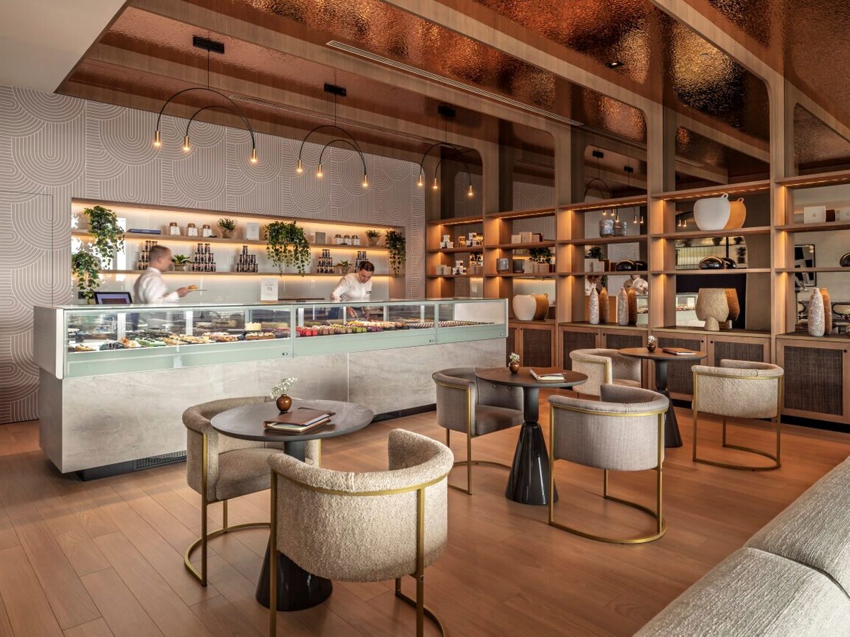 Patisserie counter with a glass cabinet of cakes and pralines, two staff behind it, product shelving along the walls and small round tables with cream bouclé armchairs.