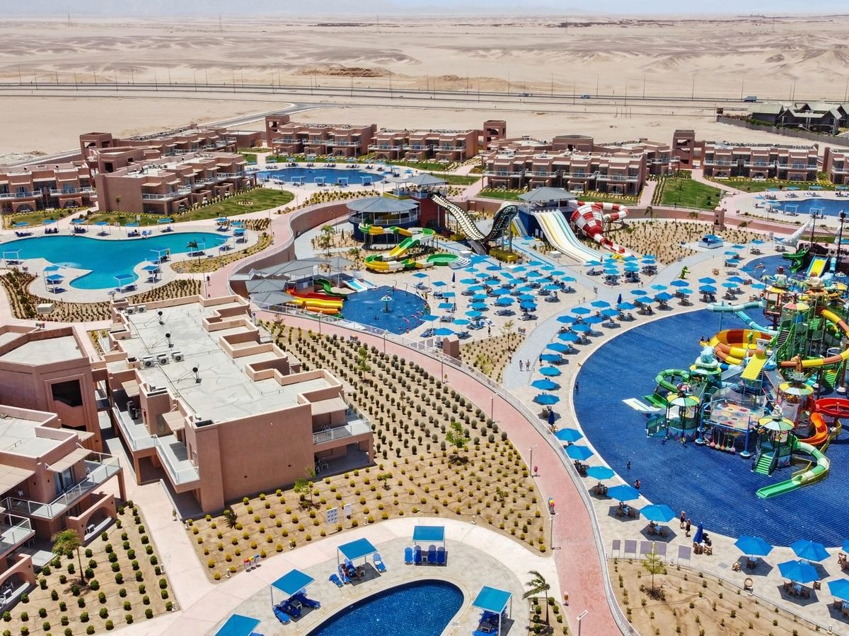 Two-storey terracotta bungalow blocks and curving blue pools laid out across a sandy site, with slides and blue parasols on one side and open desert behind.