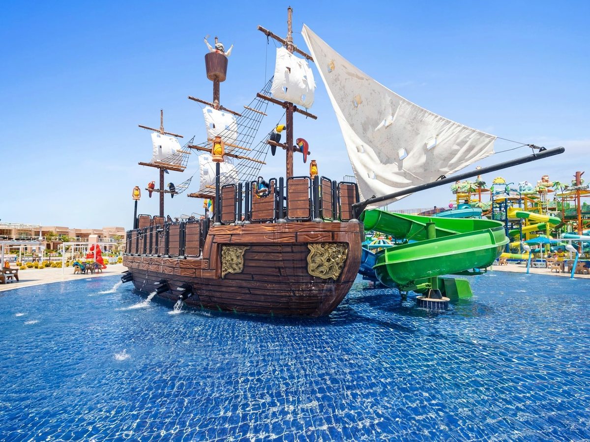 A wooden pirate ship with white sails and a green slide standing in a shallow blue splash pool, with slide towers behind.