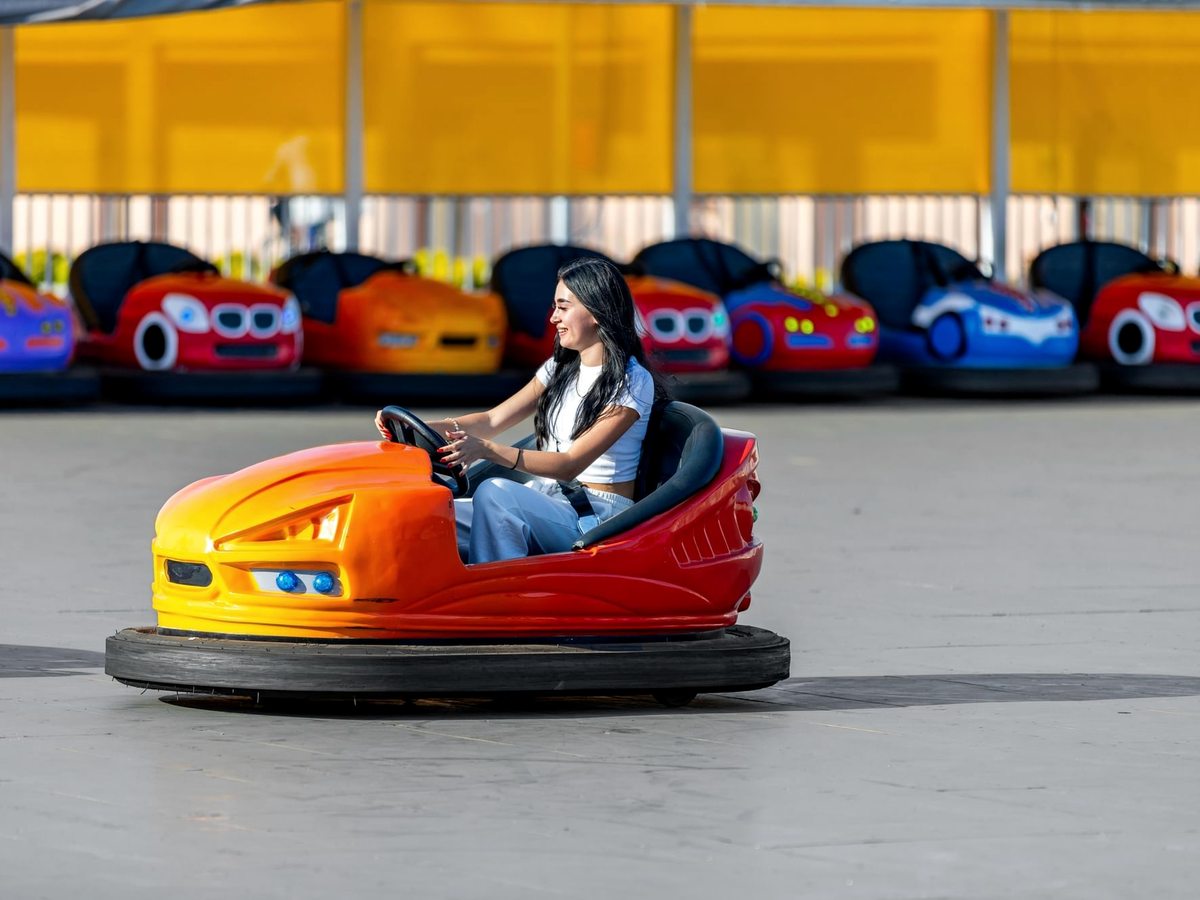 A driver in an orange bumper car on a polished floor, with blue, red and orange cars parked in a row under a yellow canopy behind.