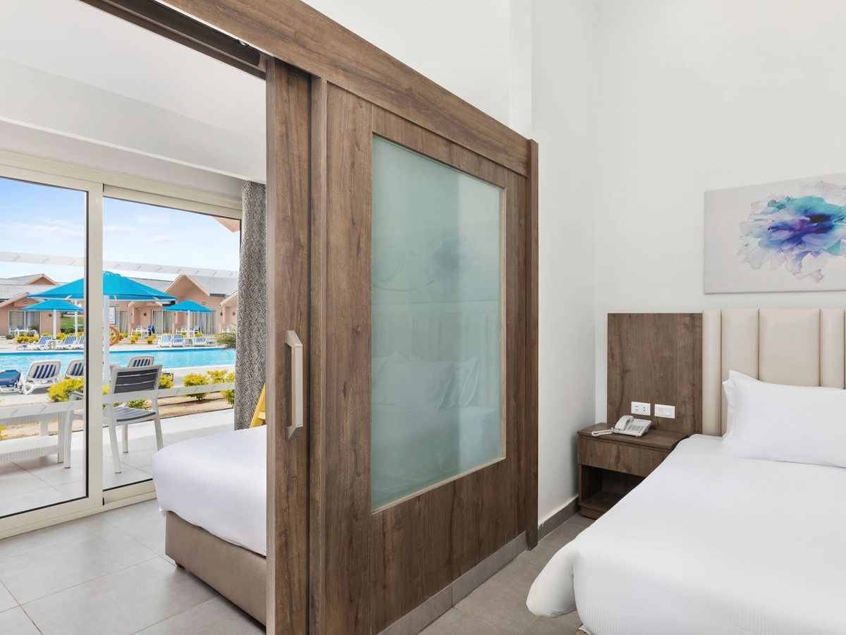 A bungalow bedroom with a cream padded headboard and a second bed beyond a sliding wooden partition, with glass doors onto a terrace and the pool outside.