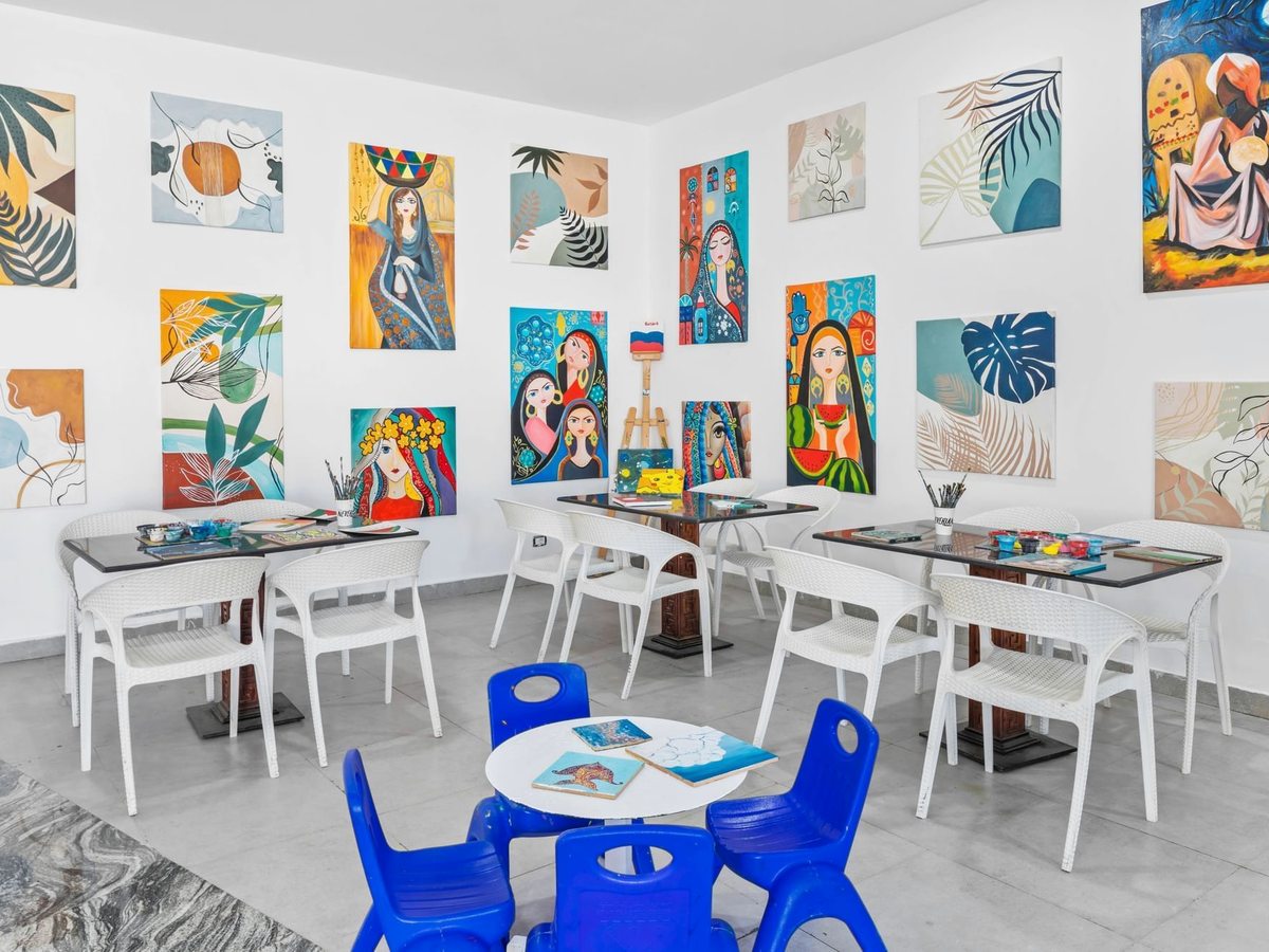 A white studio hung with painted canvases, with white tables of paint pots and brushes and small blue children's chairs in the middle.