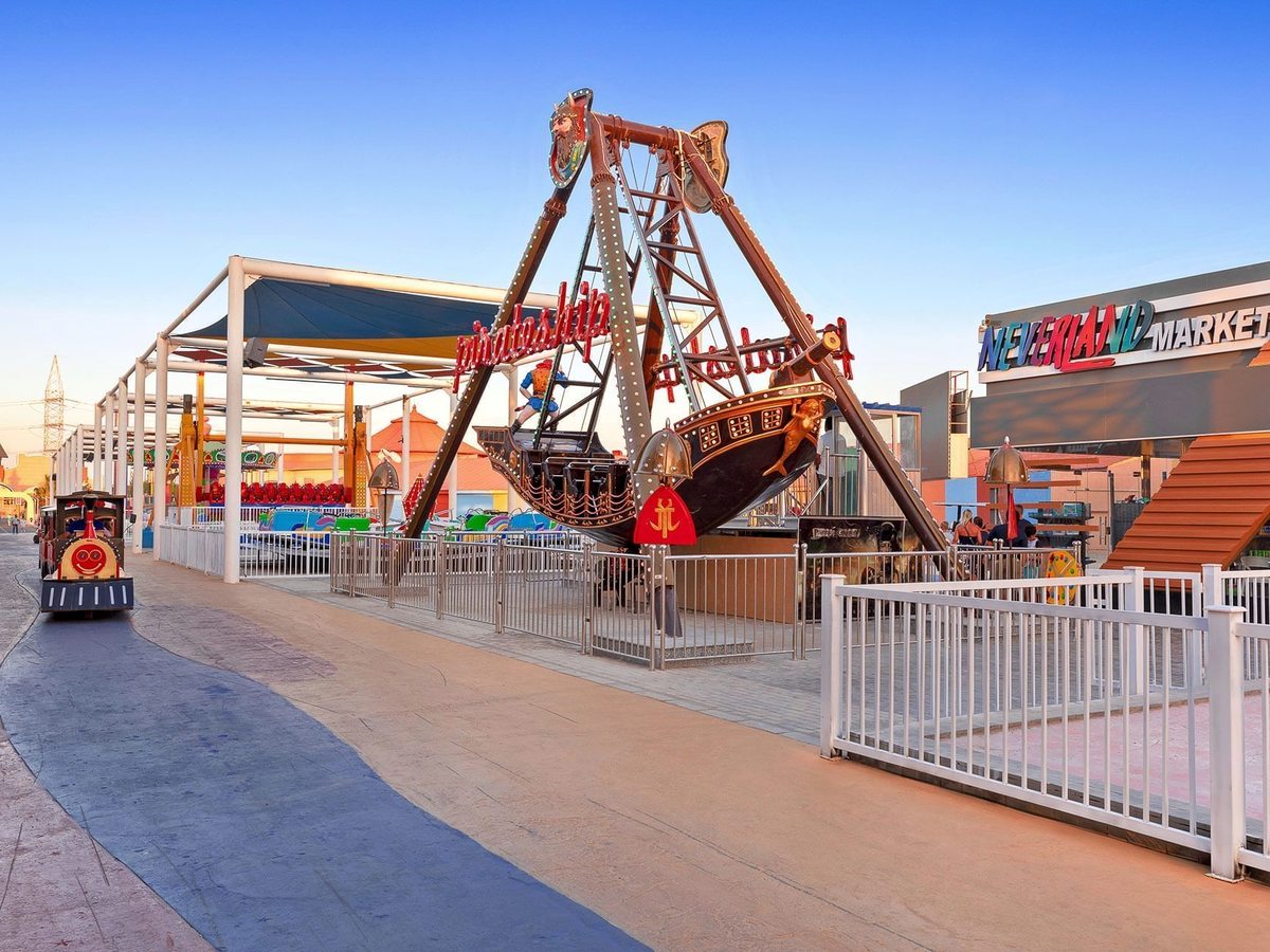 A swinging pirate ship fairground ride behind white railings, with a road train alongside and the Neverland Market building beyond.