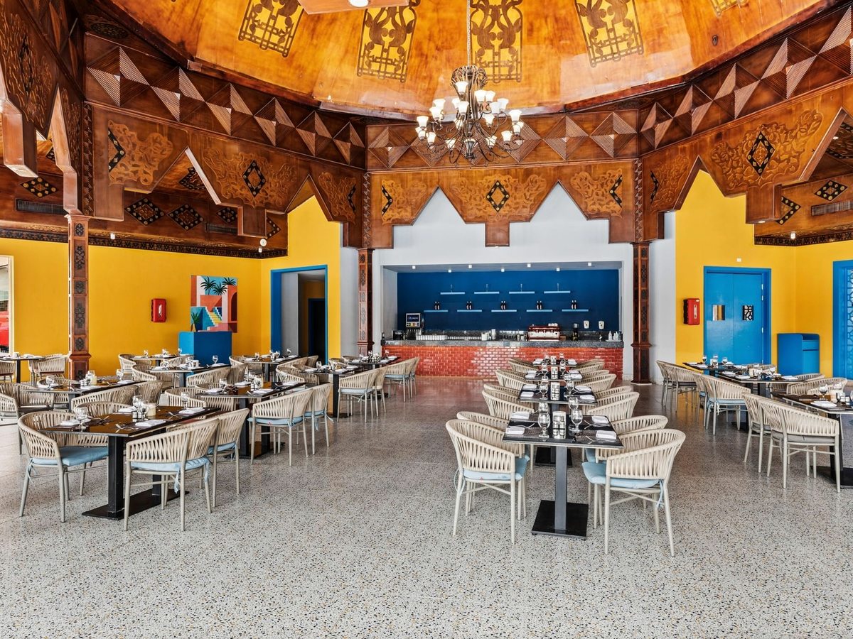 A dining room under a carved wooden dome with a chandelier, yellow walls, blue doors and laid tables with cream woven chairs.