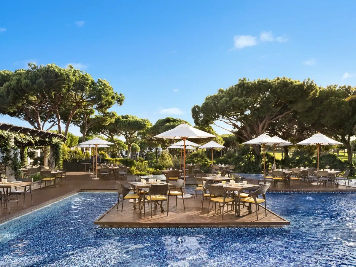 Tables and chairs under white parasols on a wooden deck set within a large pool, with umbrella pines and lawns behind.
