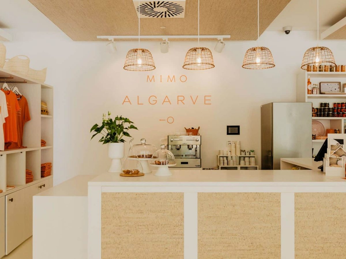 A bright white cafe counter with an espresso machine, woven pendant lights and shelves of products, under a MIMO Algarve sign.