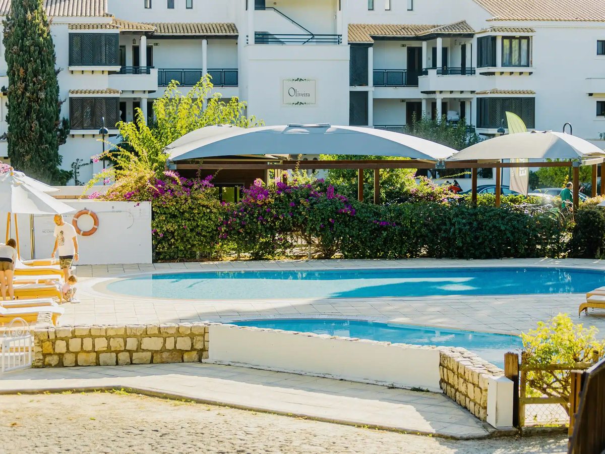 A small shallow pool separated by a low stone wall from a larger curved pool, with sun loungers and an apartment building behind.