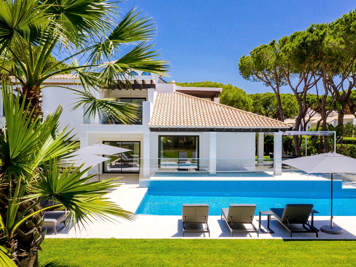 A contemporary white villa with a tiled roof and glass doors beside a private pool, with sun loungers and parasols on a lawn among palms and pines.