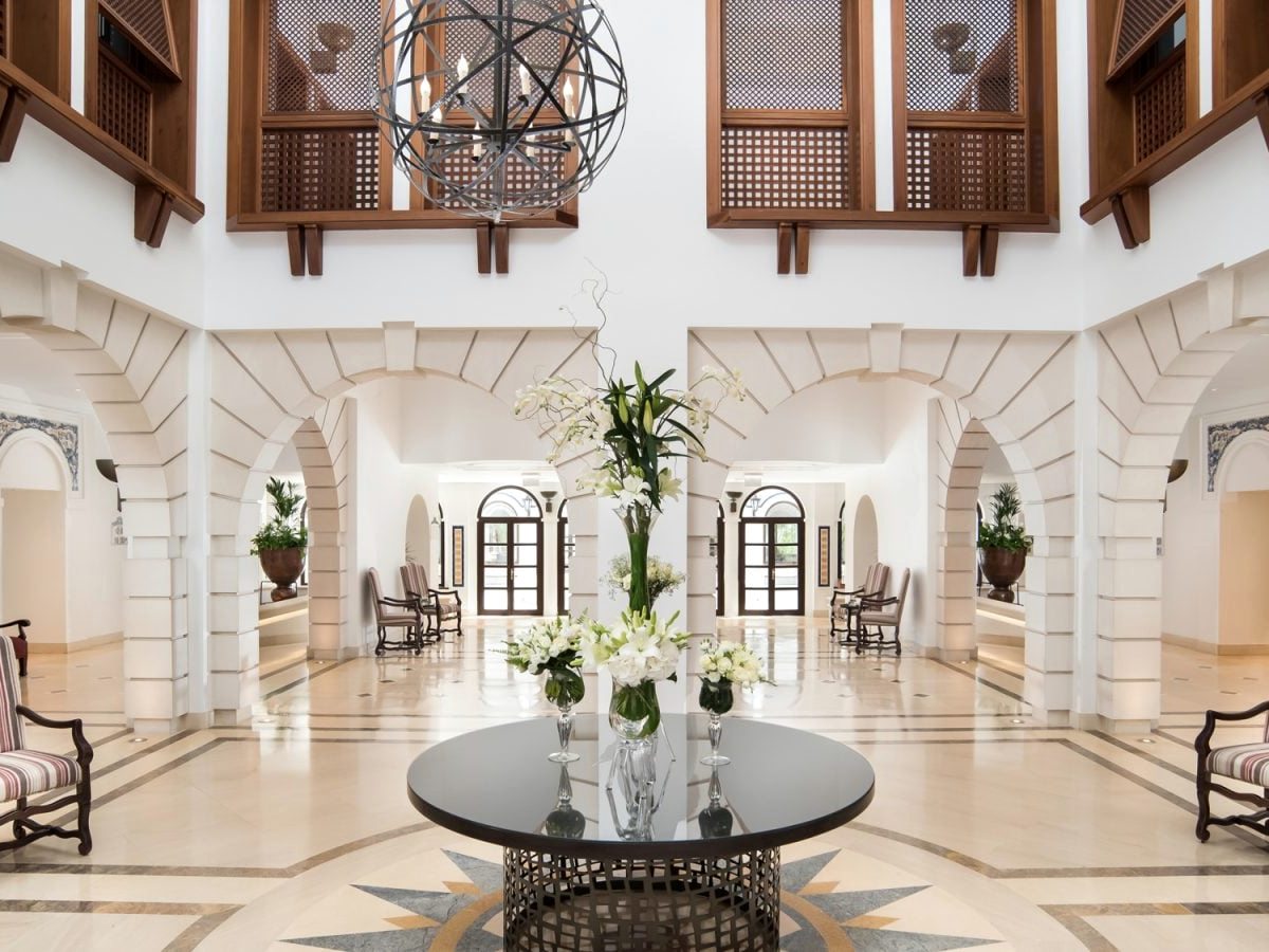 A white lobby atrium with tall arches, a lantern skylight, first-floor balconies and a round table of flowers on the tiled floor.