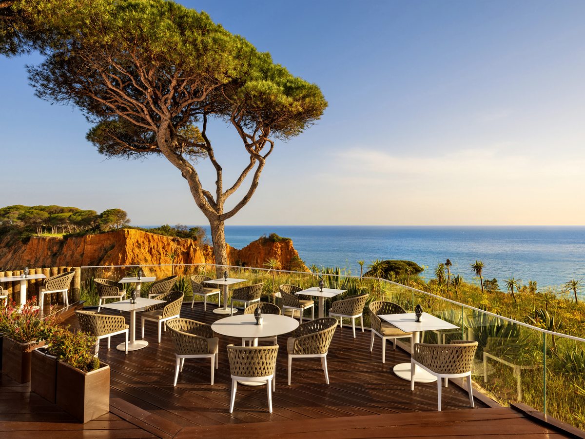 A wooden deck of round tables and woven armchairs at the cliff edge, under an umbrella pine, with red sandstone cliffs and the sea beyond.