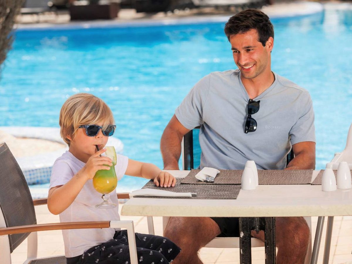 A man and a small boy with a cold drink at a table beside the pool, the blue water and poolside seating behind them.