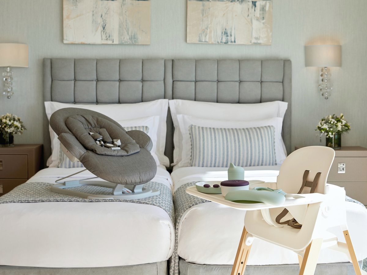 Twin room with padded grey headboards, a baby bouncer on one bed and a wooden high chair set with a beaker and children's bowls.