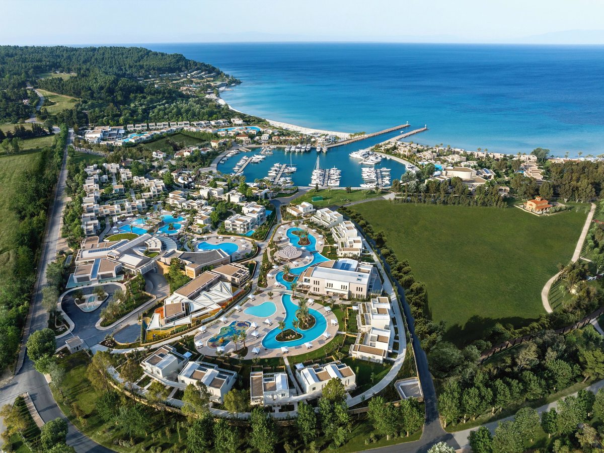 Aerial view of a low-rise white resort set among pine woods and lawns beside a yacht marina, with a sandy beach and the blue Aegean beyond.