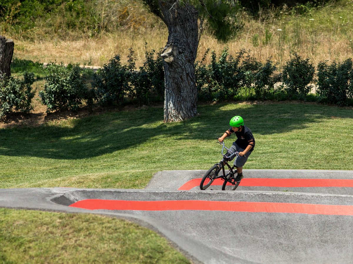 A helmeted cyclist riding over the red-marked rollers of a paved pump track, with mown grass and mature pine trees around it.