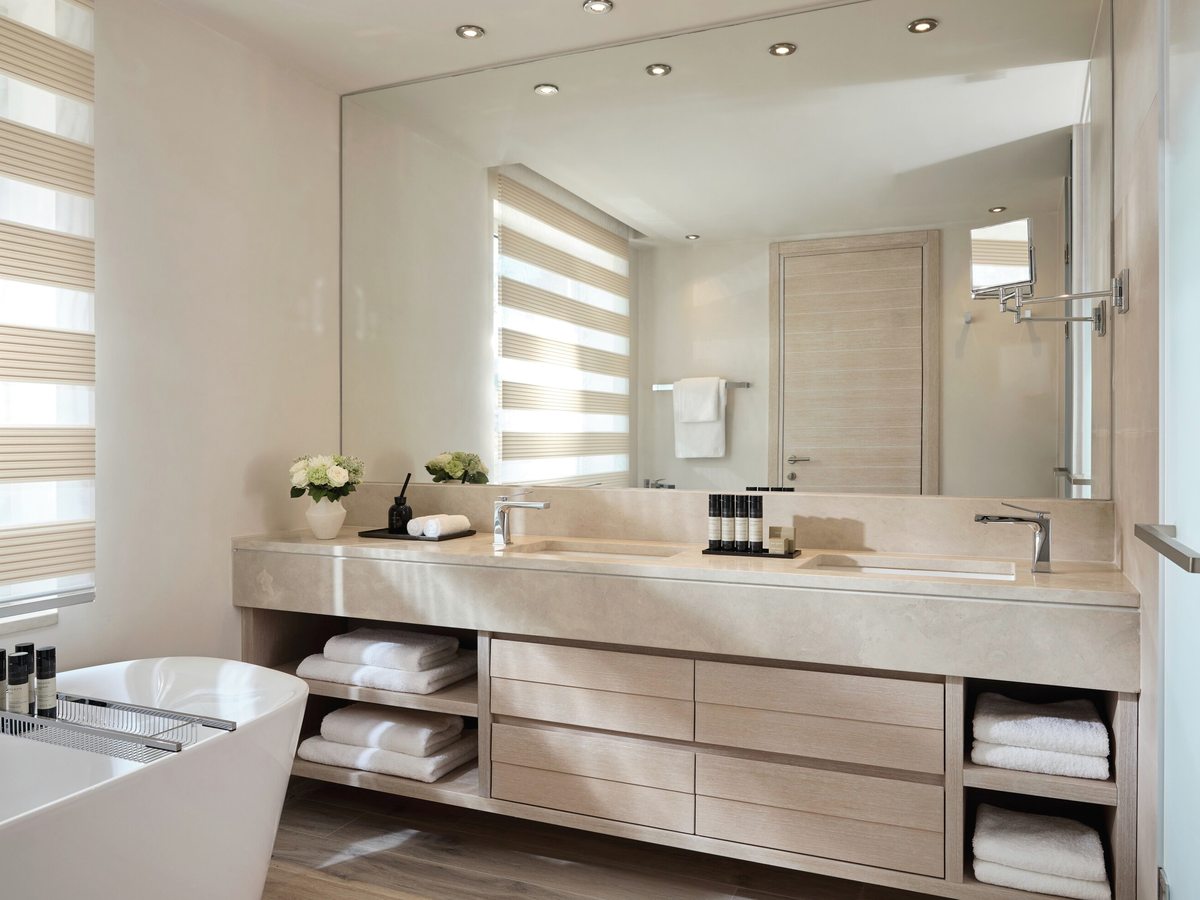 Suite bathroom with a long stone vanity and washbasin, open shelves of folded towels, a large mirror and a bath beside a shuttered window.