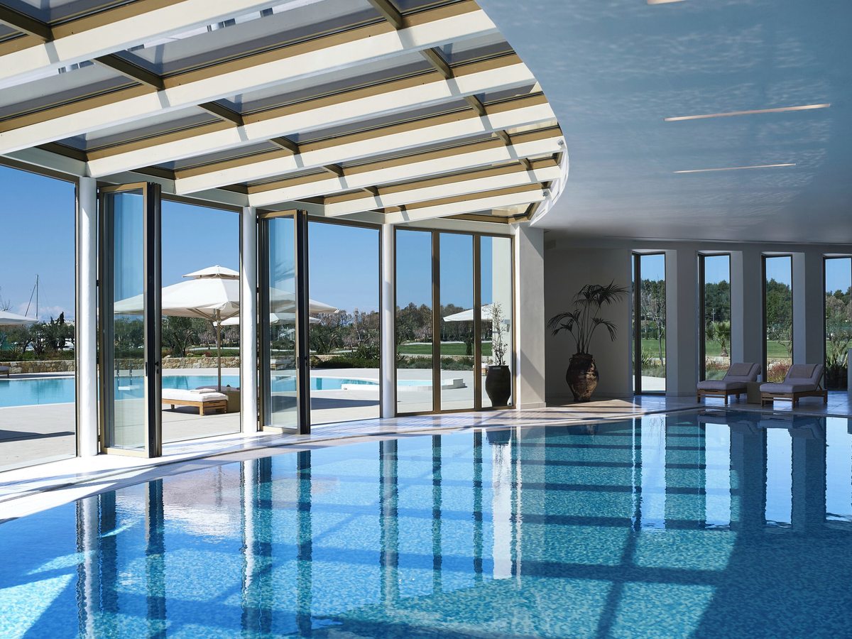 Indoor blue-tiled pool under a curved glazed roof, with folding glass doors opened onto an outdoor pool terrace and gardens.