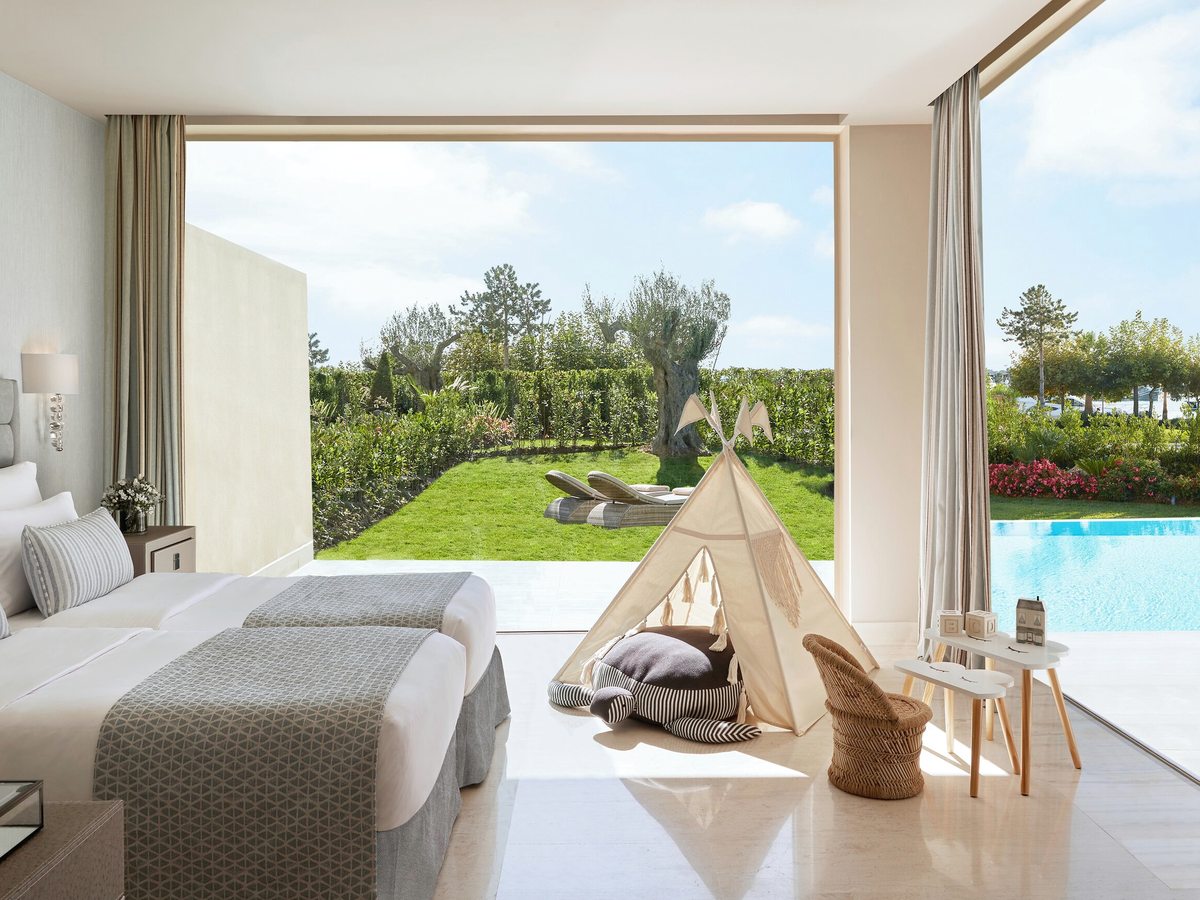 Twin room with a canvas teepee and cushions on the floor, a small child's table and stool, and glazed doors open to a lawn and pool.