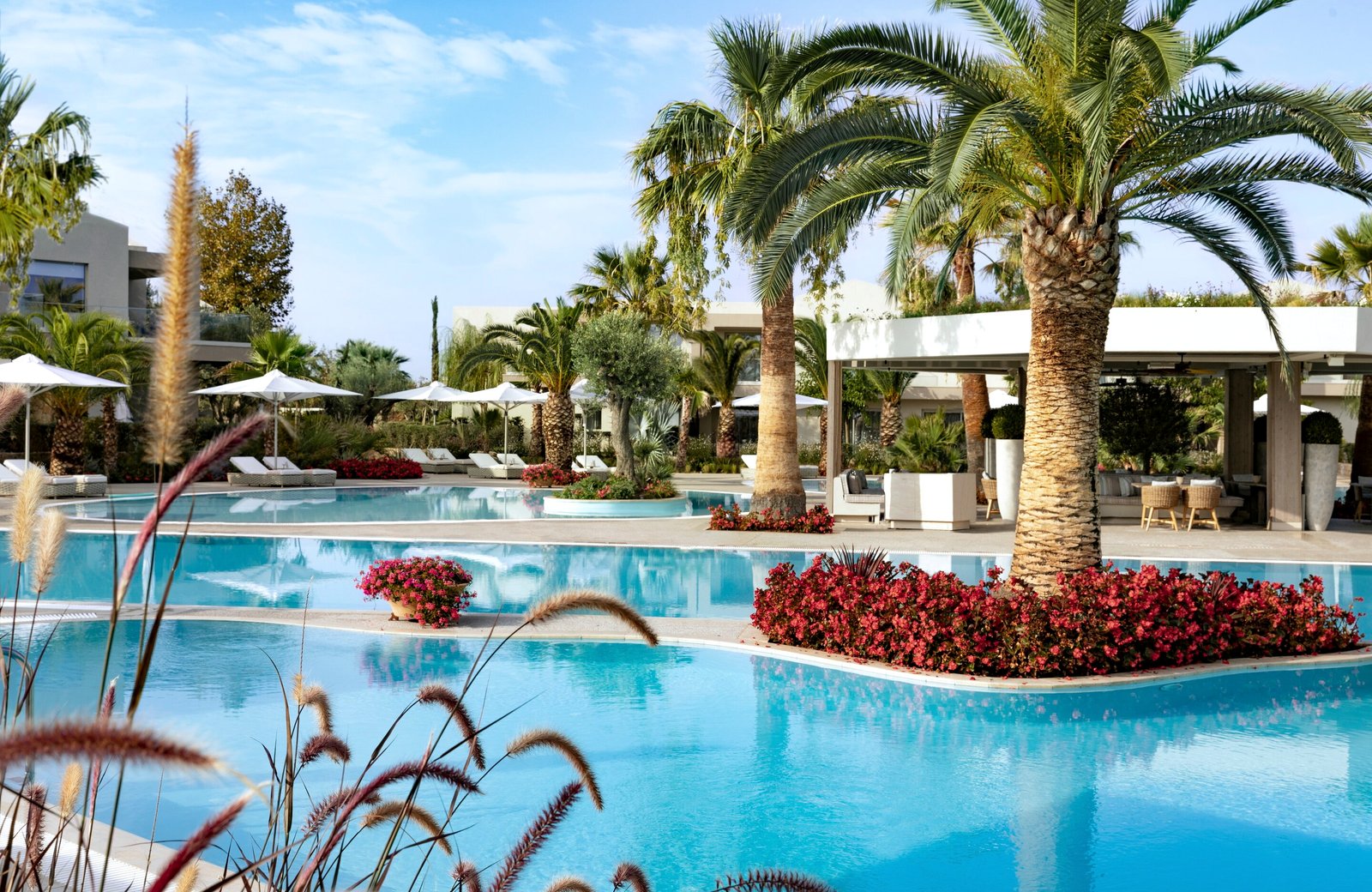 A curving turquoise lagoon pool at Porto Sani, with palms and beds of red flowers on planted islands, sun loungers under white parasols and low white buildings behind.