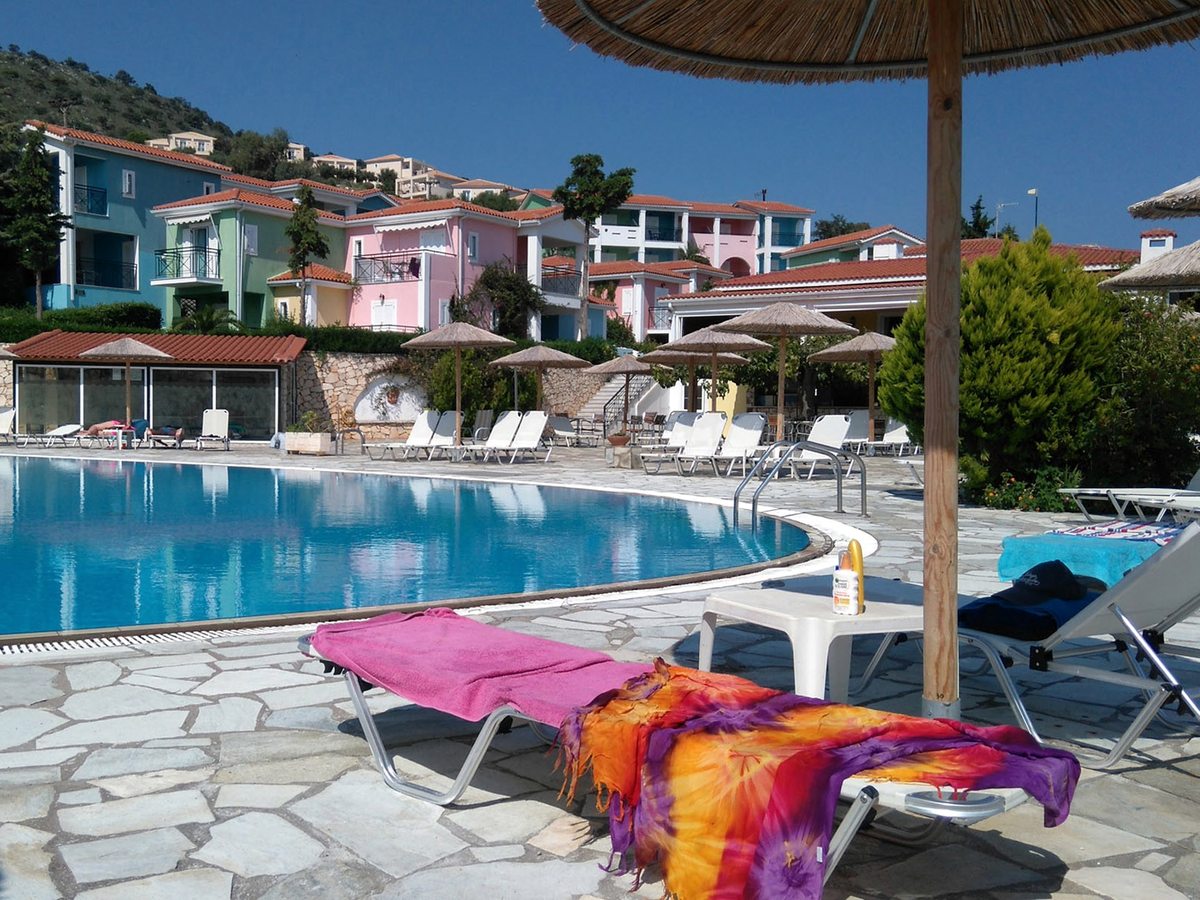 Sun loungers spread with bright towels under a thatched parasol beside the curved pool, with pastel accommodation blocks on the slope behind.