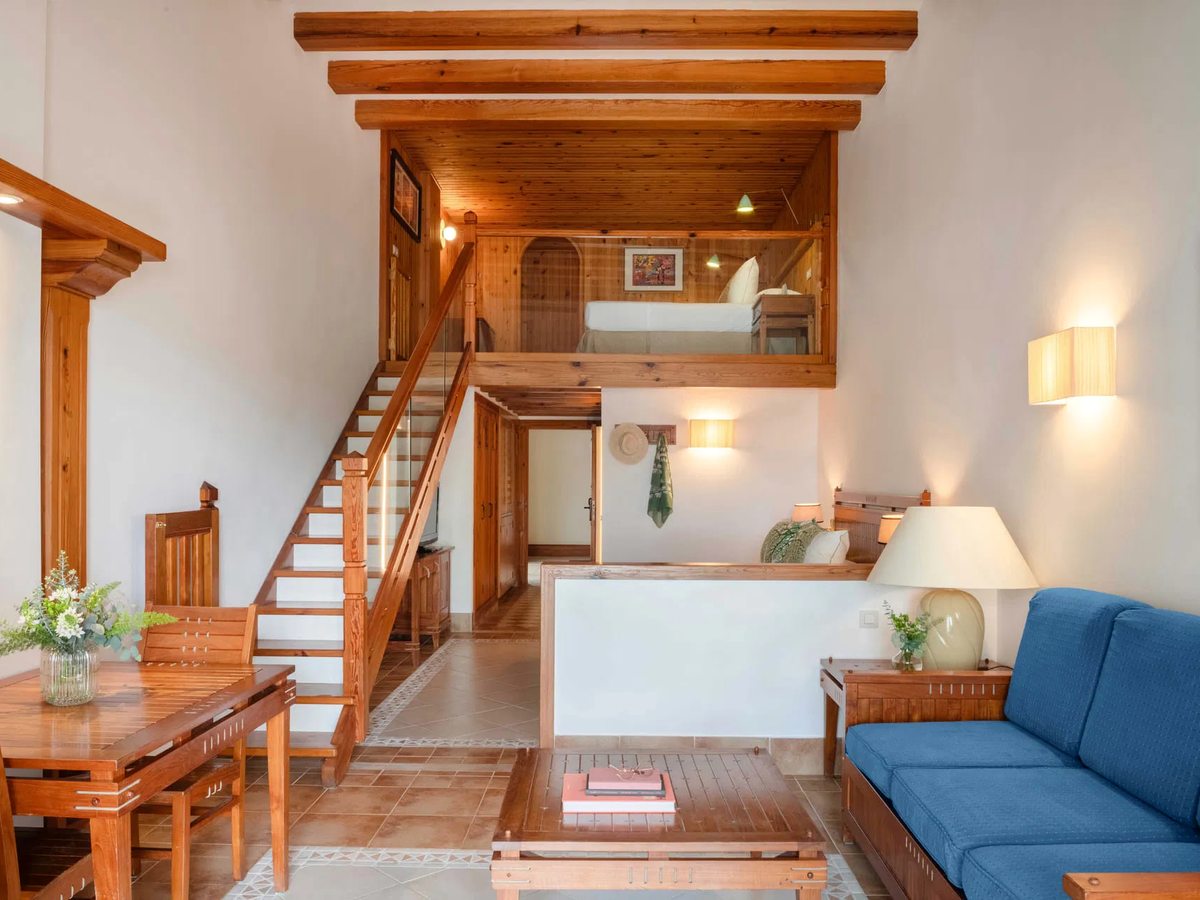 A split-level room with a wooden staircase rising to a mezzanine bedroom, and a blue sofa, coffee table and dining table on the lower floor.