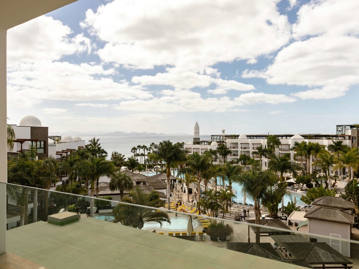 Elevated view across the resort's palm gardens and blue pools to low white buildings with domed roofs, the Atlantic and a distant island beyond.