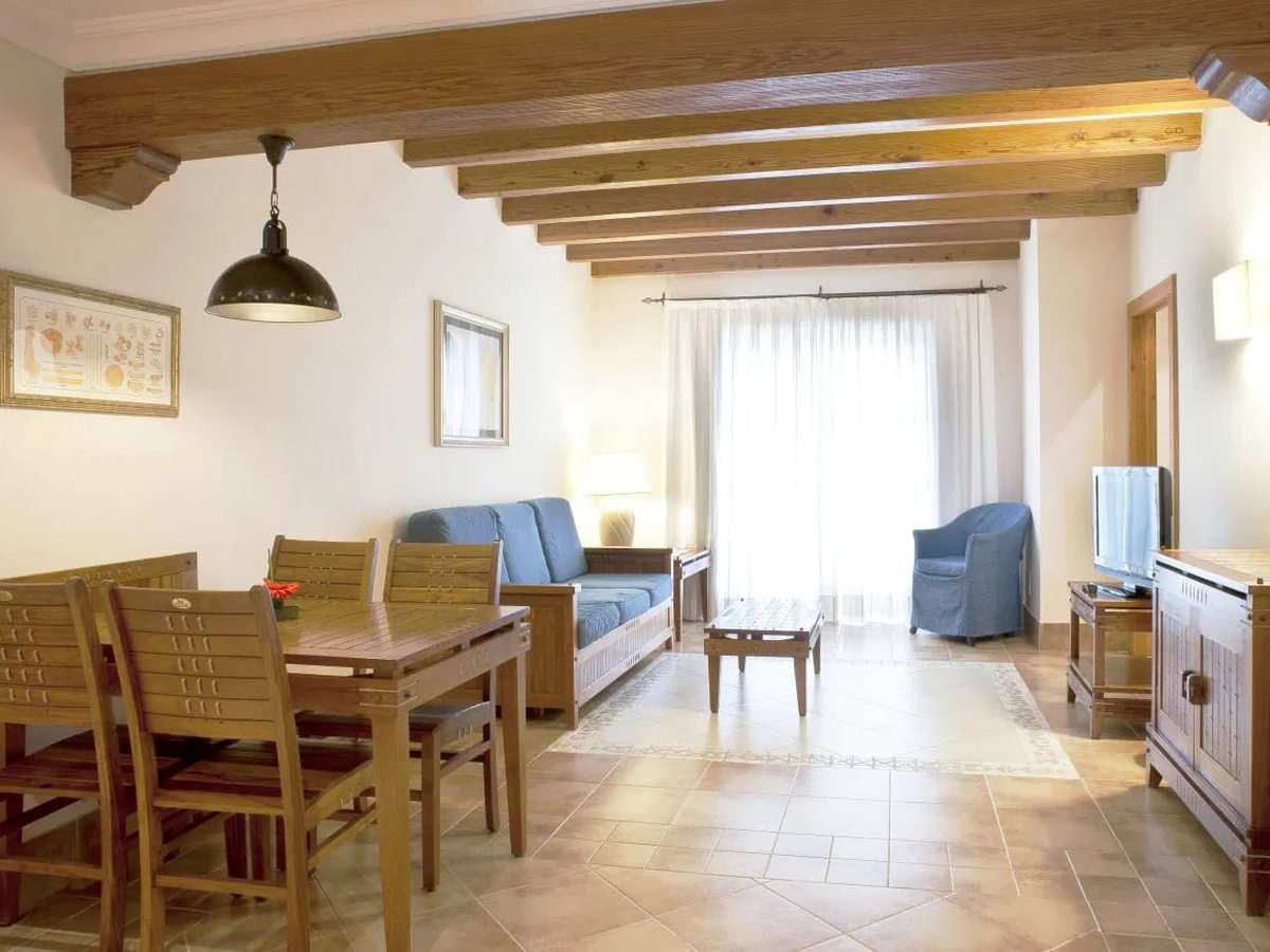 A suite living room with heavy wooden ceiling beams, a blue sofa and armchair, a wooden dining table for four and curtained doors onto a balcony.