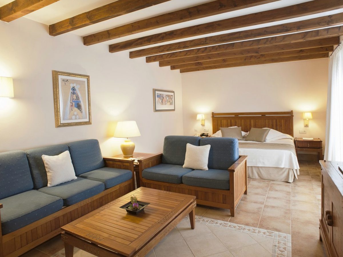 A room with exposed dark ceiling beams, two blue sofas and a wooden coffee table in the foreground and a large bed with a carved wooden headboard behind.