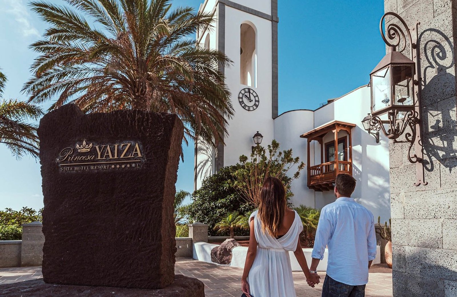 The resort's main entrance beside a tall dark stone sign reading Princesa Yaiza Suite Hotel Resort, with a date palm, a white bell tower and a wrought-iron lantern on a stone wall.