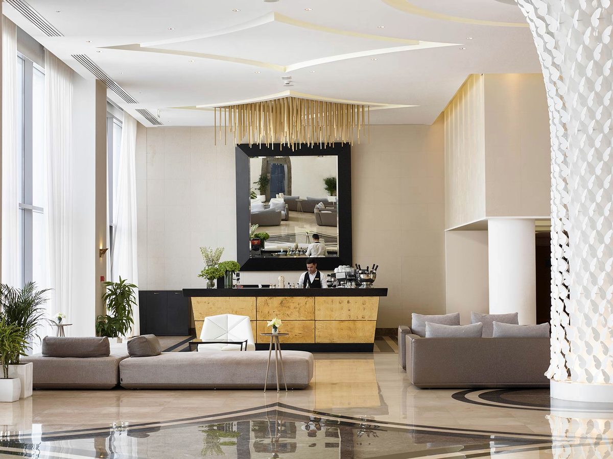 Lobby bar with a gold-fronted counter, a gold rod chandelier hanging above it and grey sofas on a polished marble floor.