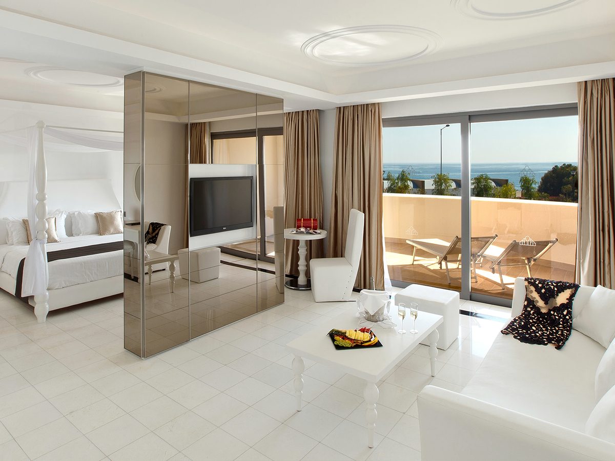 Open-plan white suite with a four-poster bed behind a mirrored divider, a white sofa and coffee table, and sliding doors to a balcony with sun loungers and a sea view.