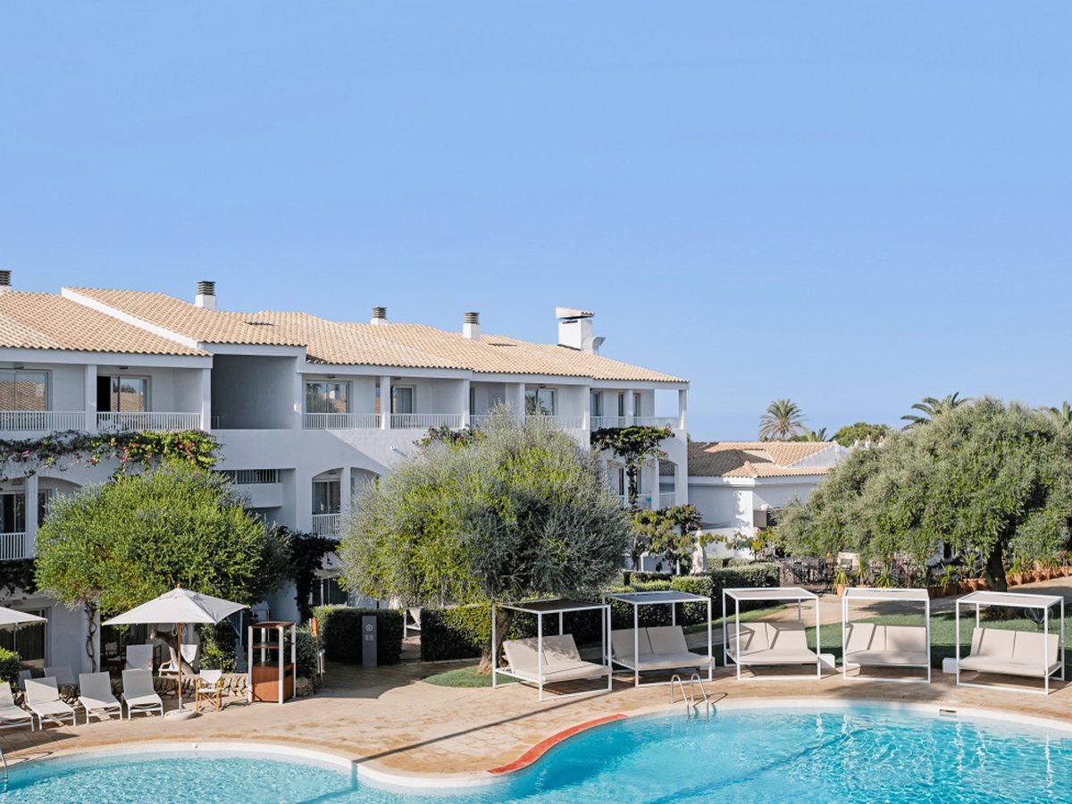 A curved outdoor pool with white four-poster daybeds, parasols and sun loungers on the terrace, in front of a white two-storey building among olive trees.
