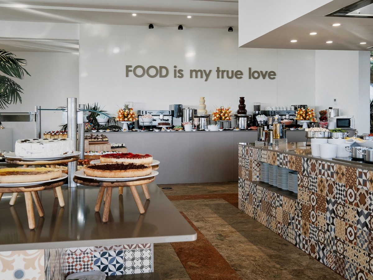 A bright buffet room with whole cakes on wooden stands, a patterned tiled serving counter, chocolate fountains and hot-drinks machines, under a wall sign reading FOOD is my true love.