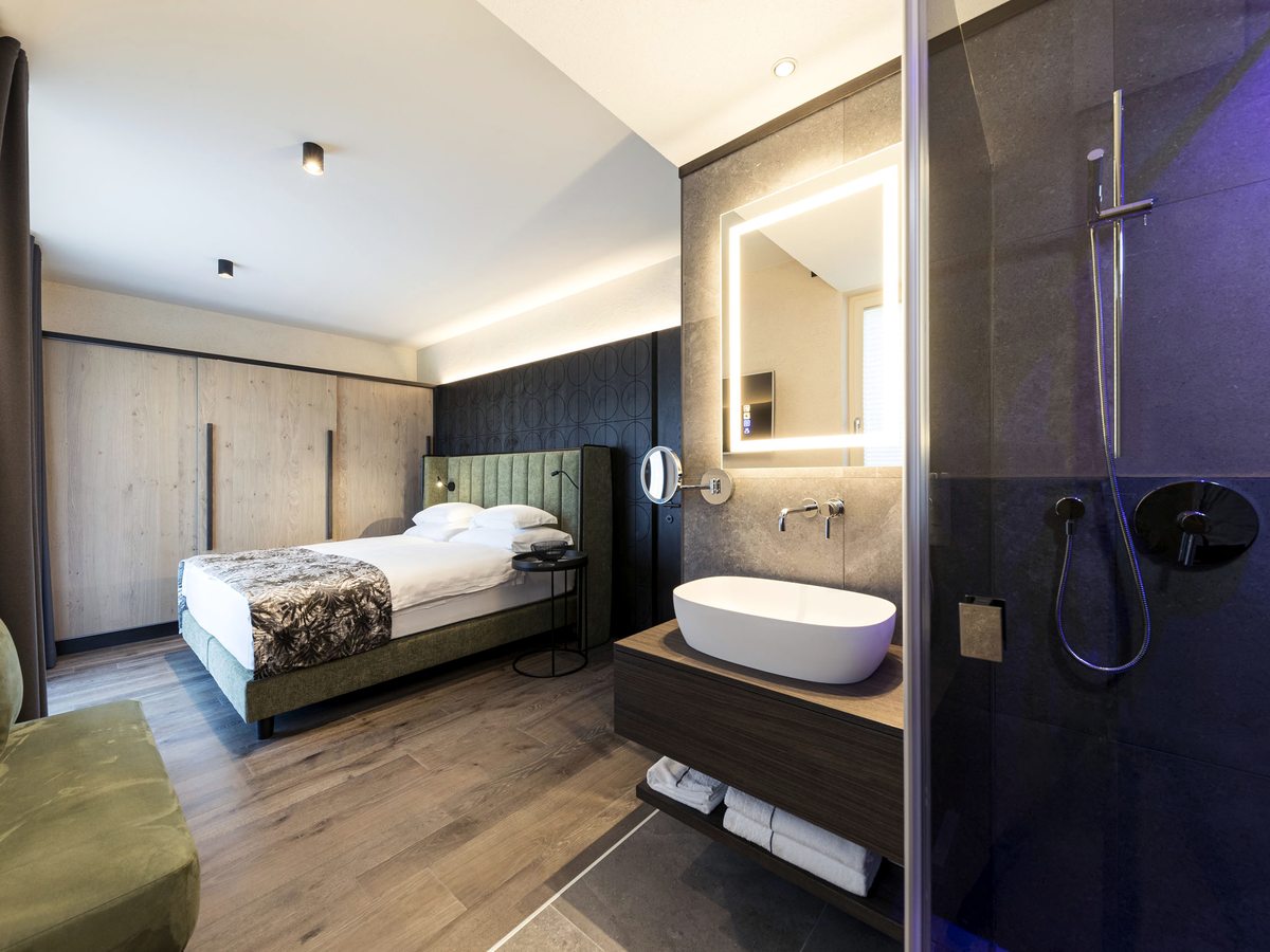 A guest room with a green upholstered bed and fitted wardrobes, and an open bathroom area with a basin on a timber vanity, an illuminated mirror and a glass-walled shower.