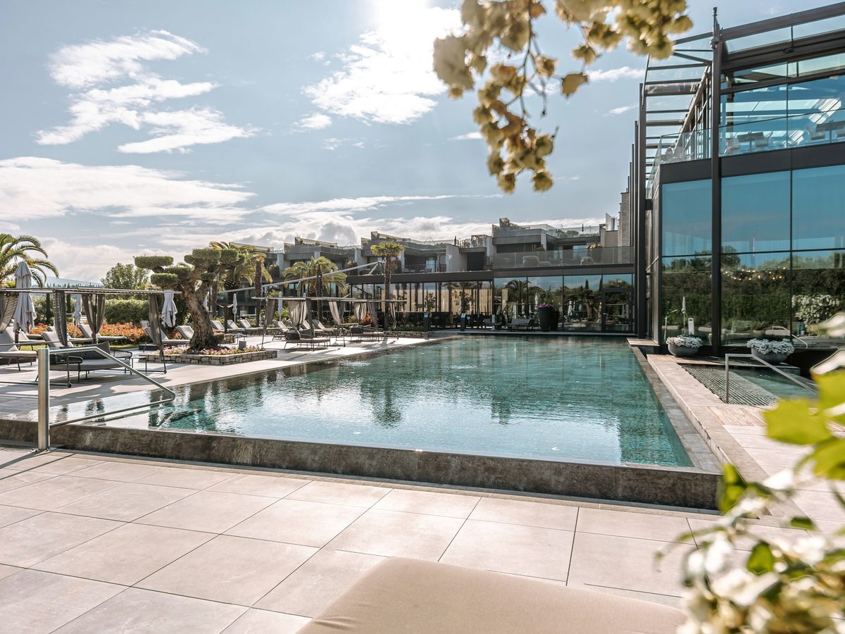 A long rectangular outdoor pool with a shallow entry shelf beside a glass pavilion, with sun loungers, cabana daybeds and palms along the tiled terrace.